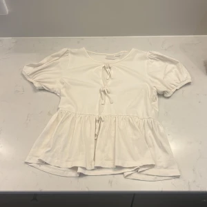 Vit blus med knyt från Gina Tricot - Säljer en söt vit blus från Gina Tricot i storlek 158/164. Blusen har puffiga korta ärmar, två knyt framtill och en volang nedtill som ger en flowy känsla. Materialet är mjuk bomull och toppen har rund halsringning. PASSAR SOM XS eftersom jag har xs och de passar mej