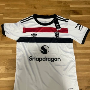 Manchester United fotbollströja Adidas - Snygg Manchester United fotbollströja från Adidas i slim fit. Tröjan är vit med svarta och röda horisontella ränder, svart krage och svarta detaljer på ärmarna. Materialet är lätt och andas, perfekt för träning. Ikoniska Adidas-ränder på axlarna och klubbmärke på bröstet. Storlek S men passar även M, aldrig använd prislappen är kvar! 