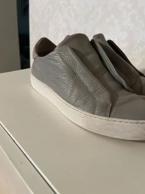 Grå sneakers från Axel Arigato - Snygga grå sneakers från Axel Arigato i mjukt skinn med vit platt sula och rund tå. Skorna har en minimalistisk design utan snörning och en kontrasterande beige mockadetalj vid hälen. Perfekta för dig som gillar stilrena och moderna sneakers. Pris kan diskuteras. Säljes i befintligt skick. 