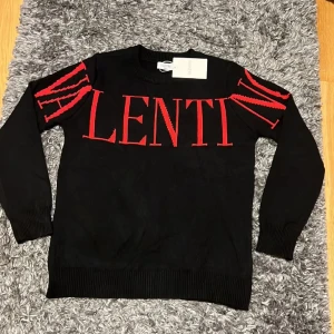 Svart Valentino tröja - Säljer en svart långärmad tröja från Valentino med stora röda bokstäver och coola röda detaljer på axlarna. Tröjan är i 100% bomull och har ribbade muddar vid ärmslut och nederkant. Perfekt för dig som vill sticka ut med en exklusiv streetstyle.
