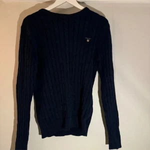 Mörkblå stickad tröja från Gant - Snygg mörkblå stickad tröja från Gant med ribbad struktur. Tröjan har långa ärmar och en diskret logga på bröstet. Perfekt för kyligare dagar.