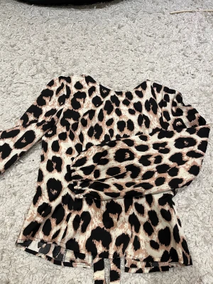 Leopardmönstrad blus med knyt från SHEIN - Säljer en snygg blus från SHEIN i storlek XS med leopardmönster i beige, svart och bruna toner. Blusen har långa ärmar med resår vid handlederna och flera knyt framtill som cool detalj. Materialet är mjukt och lätt polyester, perfekt för att sticka ut.