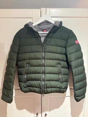 Grön pufferjacka från Colmar - Snygg mörkgrön pufferjacka från Colmar med huva och två praktiska fickor med dragkedja. Jackan har ett quiltat mönster och Colmar-logga på ärmen. Insidan är ljusgrå och jackan stängs med en rejäl dragkedja framtill. Perfekt för kalla dagar. Storleken är XSS-XS, nypris 3199 mitt pris 1299