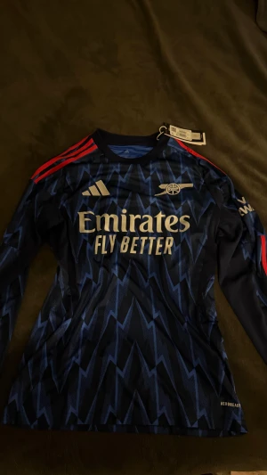 Arsenal långärmad bortatröja Adidas S - Snygg Arsenal långärmad bortatröja från Adidas i mörkblått med blixtmönster och röda ränder på axlarna. Tröjan har tryckt Emirates Fly Better-logga och Arsenal-emblem på bröstet. Tillverkad i lätt och ventilerande polyester med Aeroready-teknologi.
