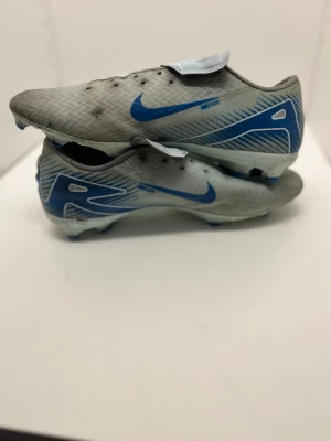 Nike grå-blå fotbollsskor - Säljer ett par gråa Nike fotbollsskor med blå detaljer och logga. Skorna har en låg profil, rund tå och platt sula med dobbar för bra grepp på planen. Ovandelen är i syntetmaterial med mönstrad yta och snörning som täcks av en skyddande flik. Perfekt för dig som älskar fotboll.