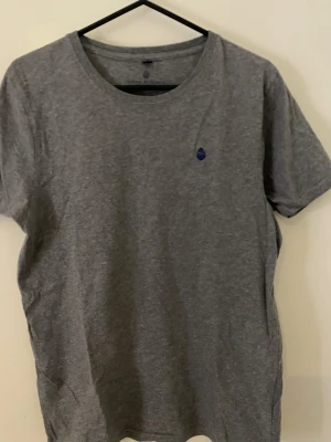 Grå t-shirt från Kordie by Kordie - Snygg grå t-shirt från Kordie by Kordie i 100% ekologisk bomull. Klassisk rund hals och korta ärmar, med en liten blå logga broderad på bröstet. Perfekt basic-plagg med skön passform och mjukt material.