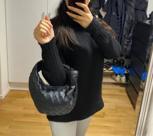 Svart flätad axelväska i skinn - Säljer en svart axelväska med snygg flätad design och rundad form. Väskan har ett kort handtag och stängs med dragkedja i guld. Tillverkad i skinn och perfekt för dig som gillar trendiga accessoarer med lite edge.