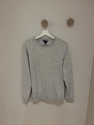Grå sweatshirt från H&M - En enkel och stilren grå sweatshirt från H&M i relaxed fit. Tröjan har rund hals, ribbade muddar vid ärmslut och nederkant samt långärmad design. Perfekt för chill dagar och passar dig som gillar en loose och bekväm stil.