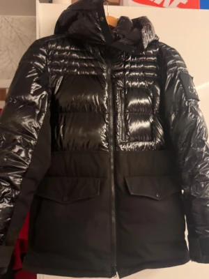 Svart pufferjacka från Moose Knuckles - Säljer en svart pufferjacka från Moose Knuckles med glansiga partier upptill och på ärmarna, samt matta partier nedtill. Jackan har huva, dragkedja framtill, stora fickor med lock och en snygg logopatch på ärmen. Perfekt för kalla dagar och riktigt stilren.