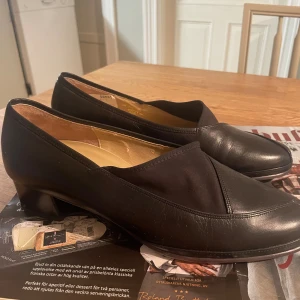 Svarta pumps från AnaNina Gabor Lady - Snygga svarta pumps från AnaNina by Gabor Lady med låg klack och rund tå. Skorna är tillverkade i äkta skinn med textildetaljer på ovansidan. Klassisk och stilren design som passar perfekt till dig som gillar en tidlös look.