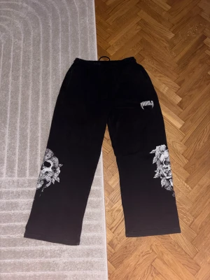 Svarta mjukisbyxor YoungLA med dödskalle - Svarta mjukisbyxor från YoungLA i storlek L med elastisk midja och snörning. Snyggt tryck med dödskalle och rosor på båda benen. Mjukt material, bred passform och bakficka. Perfekt för chill eller streetwear och gym, säljer då de var för stora och jag ryckt av lapparna, endast provade, köpta för 57€