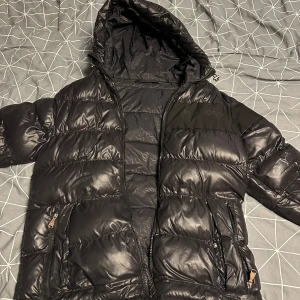 Svart pufferjacka från Alessandro Zanetti - Säljer en svart pufferjacka från Alessandro Zanetti. Jackan har en glansig finish med dragkedja och huva. Perfekt för kyliga dagar med sin vadderade design. Märkesdetaljer syns på bröstet, strl M nypris 2899 mitt pris 1049 och är ett litet hål fram på jackan men syns knappt alessandro märket fram är borttaget.