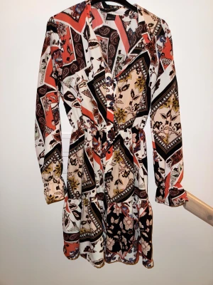 Mönstrad kortklänning med knappar från Ginatricot - Säljer en färgglad kortklänning med lång ärm och v-ringning. Klänningen har ett unikt patchworkmönster i svart, vit, beige, röd och gult med blommiga detaljer. Knappar framtill och markerad midja ger en snygg siluett. Perfekt för dig som gillar bohemisk stil.