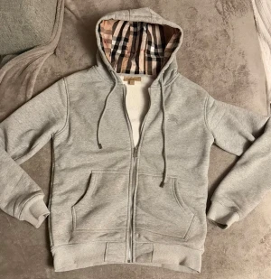 Grå Burberry hoodie med rutig huva - Snygg grå hoodie från Burberry med klassisk rutig insida i huvan och broderad logga på bröstet. Tröjan har dragkedja, snörning i huvan och två fickor framtill. Perfekt för dig som gillar stilrena och lyxiga streetwear-plagg. Skriv privat för frågor och  önskade mer detaljerade bilder ;)