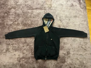 Svart Burberry hoodie med dragkedja - Svart hoodie från Burberry med klassiskt rutigt foder i huvan och guldfärgade detaljer på snörningen. Tröjan har dragkedja framtill, känguruficka och långa ärmar. Perfekt för dig som vill ha en stilren och lyxig look med ikonisk Burberry-touch.