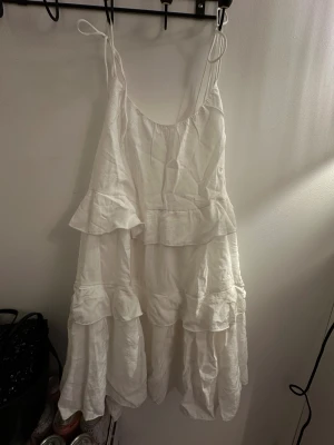 Vit volangklänning från Nelly - Supersöt vit kortklänning från Nelly med tunna axelband som knyts och flera lager volanger. Klänningen har en luftig passform och är tillverkad i mjukt bomullstyg, perfekt för varma dagar. Stilren och fräsch look med romantiska detaljer.