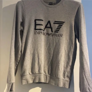 Grå EA7 Emporio Armani sweatshirt - Snygg grå sweatshirt från EA7 Emporio Armani med stort svart tryck på bröstet. Tröjan har rund hals, långa ärmar och ribbade muddar vid ärmslut och nederkant. Perfekt för dig som gillar stilrena och sportiga märkesplagg.