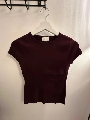Vinröd stickad topp från H&M - Säljer en vinröd stickad topp från H&M med rund hals och korta ärmar. Modellen är slim och har en enkel, stilren design utan mönster eller detaljer. Perfekt för dig som gillar basic plagg med snygg passform och mjukt material.
