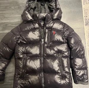 Svart pufferjacka från Polo Ralph Lauren - Snygg svart pufferjacka från Polo Ralph Lauren med röd logga på bröstet. Jackan har huva, flera dragkedjeförsedda fickor och quiltad design. Materialet är glansigt och känns lyxigt, perfekt för kalla dagar. Stängs med dragkedja framtill och har långärmade ärmar.