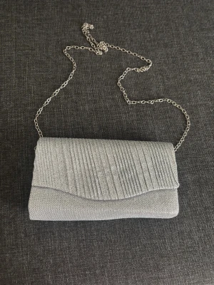Silver kuvertväska  - Snygg silverfärgad kuvertväska från Reform med glittrig yta och stilrena ränder på locket. Väskan har en tunn kedja i metall som axelrem och ett ljust innerfoder med praktisk ficka. 