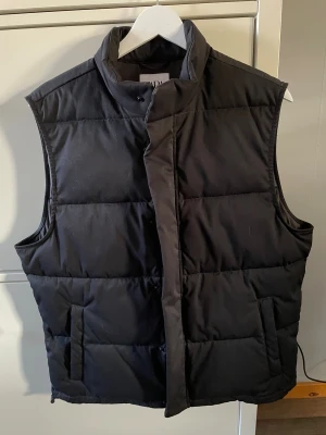 Svart quiltad dunväst från Zara - Svart dunväst från Zara i storlek M. Västen har en hög krage, quiltad design och praktiska fickor på sidorna. Stängs med tryckknappar och dragkedja.