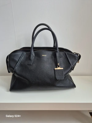 Svart handväska från DKNY i skinn - Stilren svart handväska från DKNY med guldfärgade detaljer och logotyp. Väskan har två rejäla handtag, dragkedja upptill och en snygg tag med DKNY i guld. Perfekt storlek för allt du behöver och riktigt snygg form.