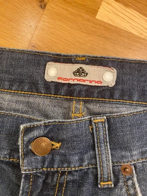 Blå bootcut jeans från Fornarina - Snygga blå jeans från Fornarina med klassisk low bootcut och slitna detaljer. Jeansen har fem fickor, gul kontrastsöm och knappstängning fram. Materialet är jeans med en tvättad look och lätt slitning vid benslut och fickor. Runt midjan är det 85cm. Länden på byxorna är 75cm. Runt låren är det 51-52cm.