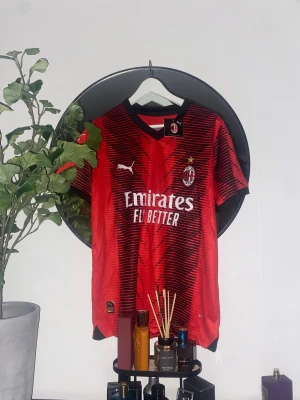 AC Milan röd matchtröja Puma - Säljer en officiell AC Milan fotbollströja från Puma i rött med svarta detaljer och grafiskt mönster. Tröjan har korta ärmar, svart krage och klubbmärke på bröstet. Materialet är lätt och andas, perfekt för träning eller match. Tryck: Emirates Fly Better och Wefox.