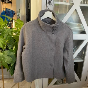 Grå kappa från Gina Tricot XS - Snygg grå kappa från Gina Tricot i storlek XS. Jackan har hög krage och fyra knappar framtill för en stilren look. Materialet är mjukt och känns som ull, perfekt för kyliga dagar. Passformen är normal och ärmarna är långa.