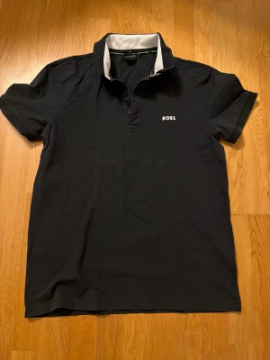 Svart Boss piké  - Svart pikétröja från Boss med vit kontrastkrage och ribbade detaljer på ärmarna. Klassisk design med korta ärmar, knappar vid halsen och Boss-logga broderad på bröstet. Tillverkad i mjuk bomull som känns skön mot huden.