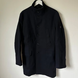 Mörkblå rock från J.Lindeberg, strl 46 - Mörkblå rock från J.Lindeberg med hög krage och knäppning framtill. Jackan har två fickor med lock och två vertikala fickor, samt snygga knappar på ärmsluten och ett bälte med knappar bak. Materialet är 75% ull och 25% polyamid, fodret är 100% viskos. Modell: Gavin Compact Melton - 4C3