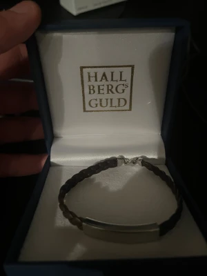 Flätat armband Hallbergs Guld - Snyggt armband från Hallbergs Guld med flätad brun rem och en stilren silverfärgad platta i metall. Perfekt accessoar för dig som gillar klassisk och enkel design med en modern twist.