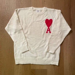 Vit stickad tröja från Ami med hjärta - Vit stickad tröja från Ami med rund hals och ribbade muddar. På bröstet finns ett stort rött hjärta och bokstaven A som ger en unik och snygg detalj. Perfekt för dig som gillar stilrena plagg med en twist.