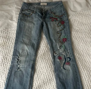 Blå jeans med broderade blommor - Snygga ljusblå jeans med broderade blommor och pärldetaljer längs ena benet. De har låg midja och bootcut, slitningar och hål på benen för en cool look. Har vissa slitningar vid hälarna. Midjemått 39cm och innerbenslängd 85cm. 