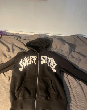 Svart hoodie från Sweet SKTBS XXS - Svart hoodie från Sweet SKTBS med stor vit grafisk text på bröstet. Tröjan har huva med snörning, dragkedja framtill och två fickor. Materialet är mjuk bomull och passformen är normal. Perfekt för dig som gillar streetwear och vill sticka ut.