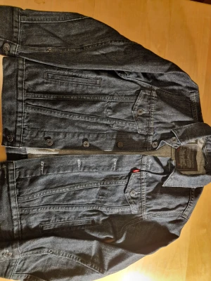 Mörkblå jeansjacka från Levi's - Snygg mörkblå jeansjacka från Levi's med klassisk design och coola detaljer som bröstfickor med knappar och den ikoniska röda Levi's-taggen. Jackan har långa ärmar, knappar framtill och är tillverkad i slitstarkt bomullsjeansmaterial. Den är i storlek M men sitter som S eller tillomed xs. Man får helt enkelt testa och se ifall den passar.
