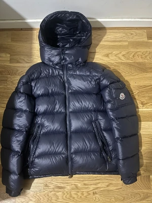Mörkblå Moncler dunjacka med huva - Säljer en mörkblå dunjacka från Moncler med stor huva och bröstficka med logotyp. Jackan har två sidofickor med dragkedja och brunt foder med serietidningsdetalj. Perfekt för kalla dagar och har en riktigt puffad look. Kvitto finns.