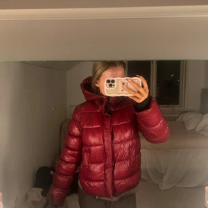 Vinröd pufferjacka med huva från Superdry - Säljer min vinröda pufferjacka från superdry. Jackan har en schysst glansig finish, stor huva och dragkedja framtill. Den är quiltad och har långa ärmar, perfekt för kalla dagar. Snygg och varm, passar dig som vill ha en varm jacka till vintern.
