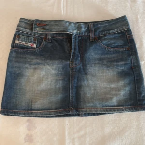 Blå jeanskjol från Diesel - Snygg blå jeanskjol från Diesel. Kjolen har dragkedja och dubbla knappar framtill samt tydlig logga på fickan. Knapp saknas dock vid gylfen men kan användas ändå då det finns ytterligare två knappar att knäppa vid sidan, se bild 1 & 3.