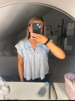 Ljusblå blus med volanger från Neo Noir - Supersöt ljusblå blus från Neo Noir med volangdetaljer längs axlarna och spetsinslag framtill. Blusen har v-ringad hals och är ärmlös med en luftig, loose passform. Perfekt för vår och sommar när du vill ha en chill och feminin look. Storlek 38 men mer som 34/36