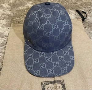 Blå Gucci keps med GG-mönster - Snygg blå keps från Gucci med det klassiska GG-mönstret över hela kepsen. Justerbar rem baktill i skinn och insida i ljus bomull. Perfekt för dig som vill ha en lyxig och trendig accessoar med tydlig designerstil.