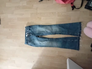 Blå bootcut jeans med broderade fickor - Säljer ett par blå bootcut jeans med slitna kanter och broderade detaljer på bakfickorna. Jeansen har klassisk femficksdesign och är tillverkade i denim. Perfekta för dig som gillar en avslappnad och trendig look.