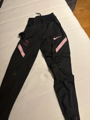 Svarta PSG träningsbyxor från Nike - Svarta träningsbyxor från Nike med rosa detaljer och PSG-logga på benet. Byxorna har snörning i midjan och smal passform med rosa ränder längs benen. Perfekta för dig som gillar fotboll och sportig stil.