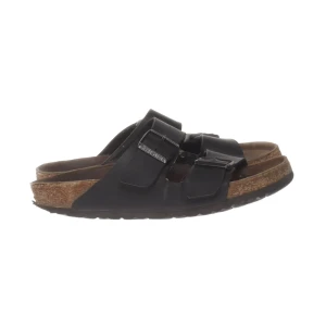 Svarta Birkenstock sandaler med spännen - Svarta sandaler från Birkenstock med två breda remmar och metallspännen. Sulan är i kork och har en ergonomisk form för extra komfort. Ovandelen är i skinn och sandalerna har en platt sula. Perfekta för varma dagar.
