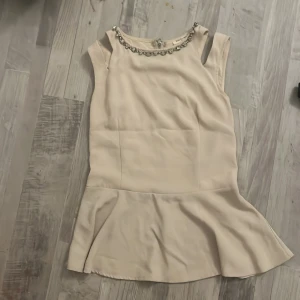 Champagnefärgad klänning Drôle de Copine - Köpt i Frankrike. Original pris 799kr Supersöt champagnefärgad lång blus från Drôle de Copine med volang nertill och ärmlös design. Klänningen har dekorativa stenar vid halsringningen och en synlig dragkedja i ryggen. Perfekt för dig som gillar en chic och feminin stil.