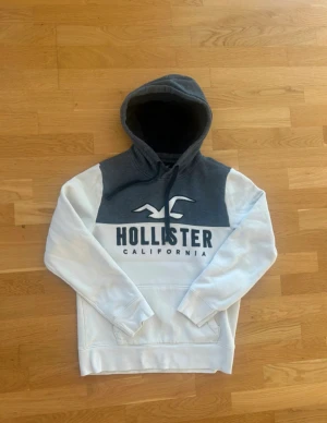 Hollister Hoodie - Säljer en snygg hoodie från Hollister i storlek S. Tröjan är vit med grått parti upptill och har stor Hollister-logga på bröstet. Utrustad med huva och snörning, samt en klassisk känguruficka framtill. Perfekt för chill dagar och streetstyle.