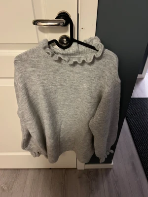 Grå stickad tröja med volangkrage - Mysig grå stickad tröja med långärmad design och en söt volangdetalj vid halsen. Perfekt för kyliga dagar och enkel att matcha med jeans eller kjol. Tröjan har en mjuk och skön känsla och passar dig som gillar stilrena plagg med en twist.