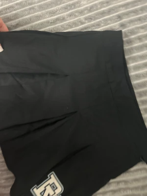 Svart kortkjol från H&M Divided HELT NY - Svart kortkjol från H&M Divided i storlek 36. Kjolen har ett stilrent utseende med veck framtill och en dekorativ patch längst ner. Den stängs med dragkedja och knapp i sidan. Materialet är mjukt och lätt, perfekt för att skapa en cool look.