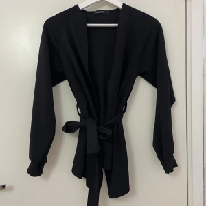 Svart omlott kavaj från Boohoo - Svart kavaj från Boohoo i omlottmodell med bälte i midjan. Jackan har långa ärmar med slits vid ärmslutet och en stilren, rak passform. Materialet är mjukt och lätt, perfekt för att ge en clean look till din outfit.