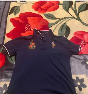  Polo Ralph Lauren Tröja - Snygg mörkblå pikétröja från Polo Ralph Lauren med klassisk broderad polospelare på bröstet och ett stilrent vapensköldsbroderi. Pikén har vit kant på krage och ärmslut samt knappar framtill. Perfekt för dig som gillar preppy stil och vill ha något som sticker ut. Om ni vill ha fler bilder skriv och buda gärna😊